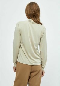 Langärmliges Oberteil in Hellbeige, aus weichem Stoff, lockere Passform, mit einem leicht drapierten Rücken und gerundetem Saum, kombiniert mit braunen Hosen.