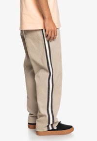 Beige kordfløyelsbukser med vertikale hvite og marineblå striper, med avslappet passform og elastisk midjebånd. Brukt med sorte joggesko.