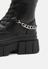 Schwarze Leder-Ankelstiefel mit einer dicken, strukturierten Sohle und silbernem Kettenakzent. Mit Schnürdesign und glatter Oberfläche.