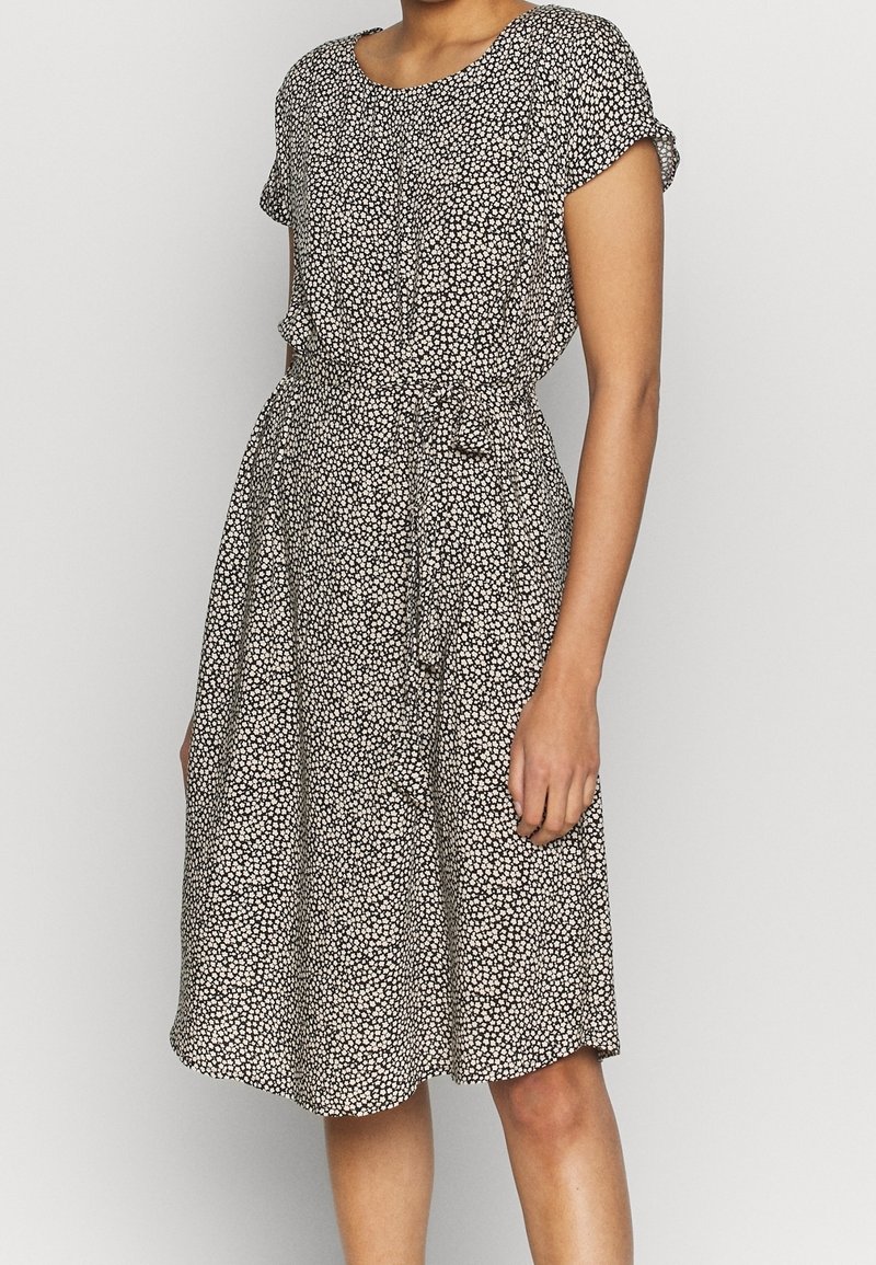 Femme portant une robe mi-longue noire et blanche à petits imprimés floraux, manches courtes et ceinture nouée à la taille.
