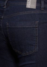 Mörkblå denimjeans med en bakficka med orange sömnad. Tyget har en slät textur och en svag lyster. Etikett synlig på midjebandet.