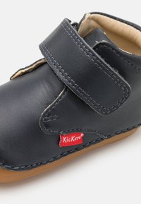 Tmavě modrá kožená bota s suchým zipem, béžovým vnitřním podšíváním a hnědou gumovou podrážkou. Na boku má červené logo Kickers.