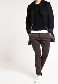 Abrigo de mezcla de lana negro sobre un sudadera negra, camiseta blanca y chinos marrones. Zapatillas blancas. Conjunto casual con diseño en capas.