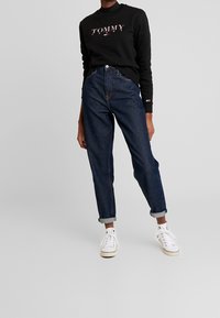 Černá mikina s logem "TOMMY JEANS" v růžové barvě, kombinovaná s tmavě modrými džíny s oholenými manžetami a bílými teniskami. Jednoduchý, volnočasový outfit.