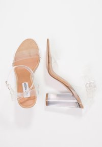 Paire de sandales beiges avec des brides transparentes à la cheville et des talons blocs, une sandale vue de dessus et l'autre de profil.