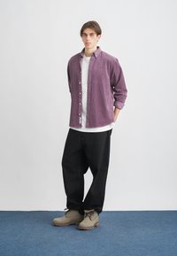 Fialová kordová košeľa na gombíky, pod ňou biela tričko, čierne voľné nohavice a béžové topánky s textúrovanou podrážkou, stojaci na modrom koberci.