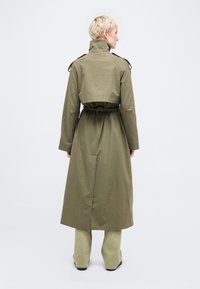 Άτομο με κοντά ξανθά μαλλιά που φοράει μακρύ χακί παλτό, ανοιχτό πράσινο παντελόνι και μαύρα παπούτσια, στέκεται μπροστά σε λευκό φόντο.