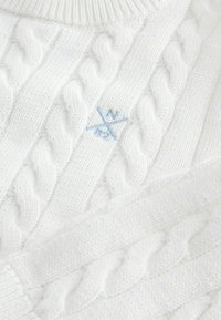 Fehér kötött pulóver, textúrázott mintákkal, középen világoskék hímzett logóval. Simaság jellemzi és bordás nyakkivágással rendelkezik.