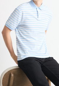 Polo shirt azzurro chiaro con strisce bianche, maniche corte e colletto morbido, con un piccolo logo sul petto sinistro. Abbinato a pantaloni scuri.