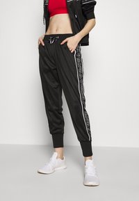 Zwarte sportieve joggingsbroek met een tapse pasvorm, voorzien van witte zijstrepen en een elastische tailleband. Gepaard met witte sneakers.