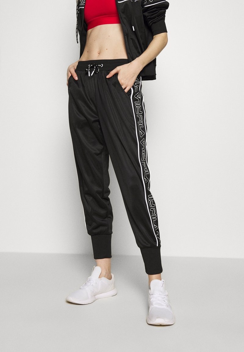 Zwarte sportieve joggingsbroek met een tapse pasvorm, voorzien van witte zijstrepen en een elastische tailleband. Gepaard met witte sneakers.