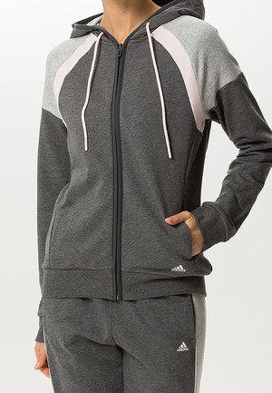 Kobieta ubrana w ciemnoszary dres Adidas z jasnoszarymi i bladoróżowymi akcentami, stojąca z jedną ręką w kieszeni kurtki.