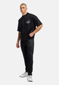 Sort kortærmet t-shirt med en dekorativ broderet design, parret med sorte jeans med diskrete mønstre, og hvide sneakers.