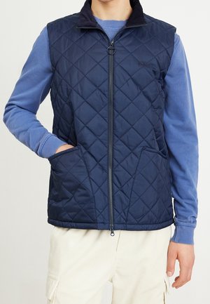 Person trägt eine marineblaue gesteppte ärmellose Weste über einem hellblauen Langarmhemd, mit offwhitefarbener Hose, steht mit einer Hand in der Tasche.