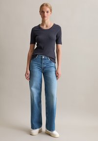 Marineblauwe ribgebreide t-shirt met korte mouwen, gecombineerd met lichtblauwe wijde jeans. Witte sneakers maken de outfit af. Neutrale achtergrond.