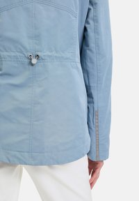 Helle blaue Jacke mit glatter Textur, ausgestattet mit einem Kordelzug in der Taille, Nahtdetails und einem reflektierenden Akzent am Ärmel.