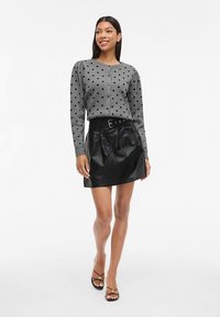 Grå genser med polkaprikkmønster og knappestenging, kombinert med et svart midjebelte i kunstlær skjørt, og svarte sandaler med stropper på en ensfarget bakgrunn.