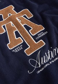 Marineblaue Fleece-Stoff mit strukturierten braunen Buchstaben "ATX" und weißen Akzenten. Enthält die eingestickten Wörter "Austin" und "US-Bundesstaat Texas".