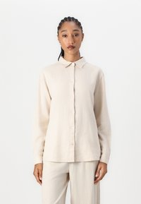Beige Langarmhemd mit Knopfleiste, Kragen und lockerer Passform. Aus strukturiertem Stoff gefertigt, kombiniert mit passenden hellbeigen Hosen.