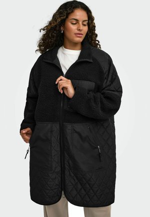 Kvinde med krøllet hår iført en lang sort quiltet og fleece lynlåsjakke over en hvid top og lyse bukser.