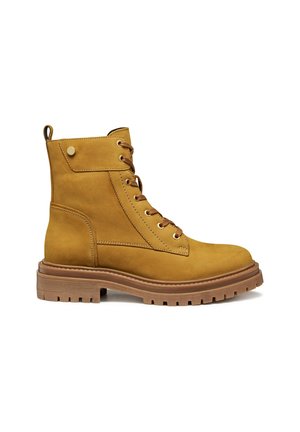 Bottines à cheville en suede beige avec un laçage sur le devant, bout rond et semelle en caoutchouc texturée. Présente des coutures décoratives et des accents de rivets en métal.