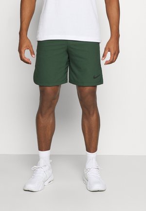 Mężczyzna w ciemnozielonych szortach Nike, białych skarpetkach, białych butach sportowych i białej koszulce, stojący na jednolitym tle.