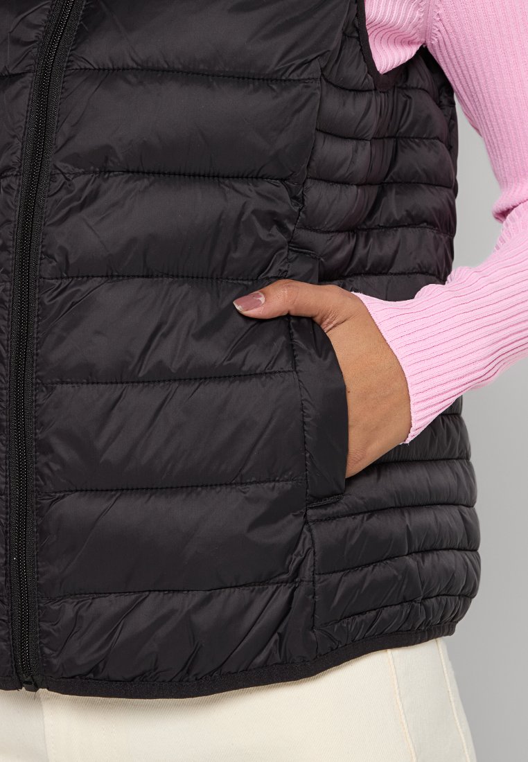 Schwarze Steppweste mit horizontalem Quiltmuster, ausgestattet mit einem Reißverschluss und seitlichen Taschen. Tragen über einem strukturierten, rosa langärmeligen Oberteil.