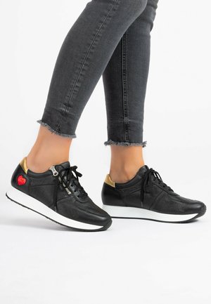 Black leather sneakers with gold accents, textured upper, white sole, and decorative red heart patch. Worn with frayed hem jeans.