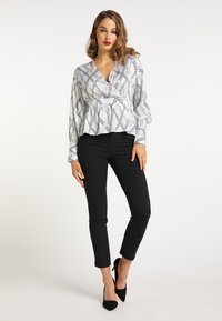 Weiße Bluse mit Kettenmuster, Peplum-Saum und langen Ärmeln, kombiniert mit schwarzen, eng anliegenden Hosen und schwarzen spitzen High Heels.