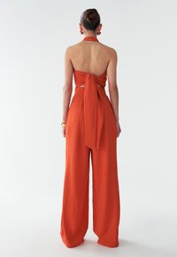 Narančasti dvodijelni outfit koji se sastoji od topa s halter vratom s vezanjem na leđima i širokih hlača od teksturirane tkanine. Glatka završna obrada, minimalno detaljiranje.