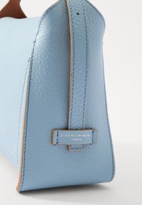 Borsa a tracolla in pelle azzurro chiaro con texture liscia, caratterizzata da un accento color cammello e un marchio impresso su un'etichetta in pelle. Forma arrotondata e dettagli di cucitura.