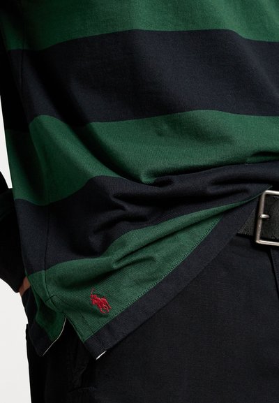 Groen en marineblauw gestreepte lange mouwen shirt met een rode geborduurde logo aan de zoom, gecombineerd met donkere broeken en een zilveren riemgesp.