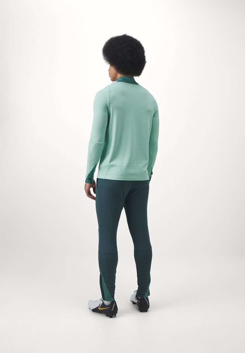 Haut à manches longues vert menthe avec texture côtelée, associé à un pantalon ajusté vert foncé avec fentes latérales. Chaussures de sport blanches et noires.