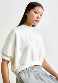 Sweatshirt creme com um decote alto, logótipo em relevo "NIKE 1972" e mangas curtas e bufantes. Combinado com calças de moletão cinzentas com cintura elástica.