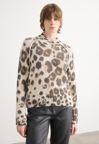 Pull en imprimé léopard dans des tons beige et marron, avec des poignets et un ourlet côtelés, col rond avec un détail zippé. Porté avec un pantalon noir.
