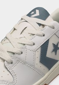 Άσπρο δερμάτινο αθλητικό παπούτσι με κρεμ κορδόνια και μπλε λογότυπο αστεριού στο πλάι και στη γλώσσα, που δείχνει λεπτομερή ραφές και υφή.