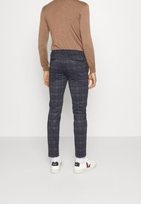 Op maat gemaakte grijs geruite broek met een slim fit en een gladde stof. Gecombineerd met een bruin langslavend shirt en witte sneakers met rode accenten.