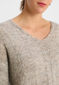 Pull en tricot beige clair avec un col en V, un motif torsadé texturé et des détails en maille ouverte sur les manches. Texture douce et duveteuse.