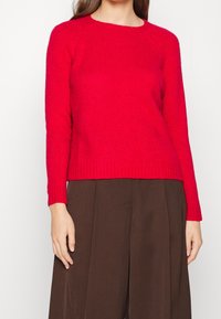 Punainen villainen neulepusero, jossa pitkät hihat, pyöreä pääntie ja resorireuna; pehmeä tekstuuri ja rento istuvuus; yhdistetty ruskeisiin leveälahkeisiin housuihin.