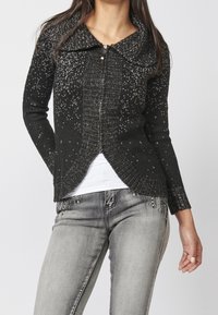 Black knit cardigan with a textured pattern, featuring a wide collar and asymmetrical hem. Zipper closure, long sleeves, and slight sparkle detailing.