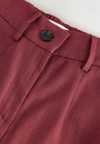 Primer plano de la cintura de pantalones granate con botón negro, trabillas para cinturón y parte trasera elástica parcialmente fruncida.