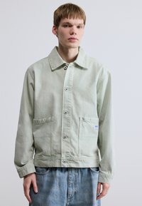 Ανοιχτό πράσινο βαμβακερό μπουφάν με δομημένο γιακά, εμπρόσθιες τσέπες και κλείσιμο με κουμπιά. Το σύνολο ολοκληρώνεται με ξεθωριασμένο μπλε τζιν παντελόνι.