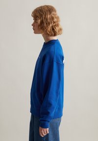 Blauwe sweatshirt met ronde hals, lange mouwen, ribgebreide manchetten en zoom. Heeft een logo op de linkermouw. Draagt met een hoge taille denim.