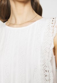 Vitt klänning med texturerad tyg och vertikala ränder samt intrikat spetsdekoration på ärmarna. Guld kedja synlig vid halsringningen.