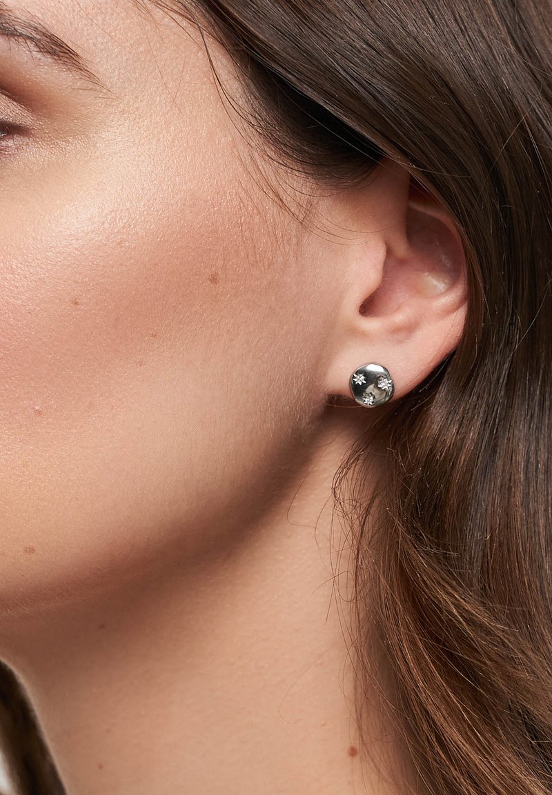 Sølvfarvede øreringe med studs, der har et sfærisk design og tre små krystalaccentuer. Glat metallisk finish med en subtil refleksiv kvalitet.