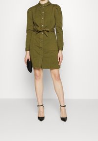 Abito camicia verde oliva con maniche lunghe, dettagli arricciati sulle spalle, cintura in tessuto e due tasche frontali. Clutch nera e décolleté a punta.
