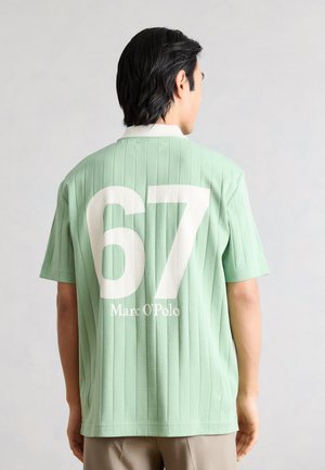 Άνδρας με μαύρα μαλλιά, που φορά ανοιχτοπράσινο κοντομάνικο πουκάμισο με κάθετες ρίγες και μεγάλο λευκό κείμενο "67 Marc O'Polo" στην πλάτη.