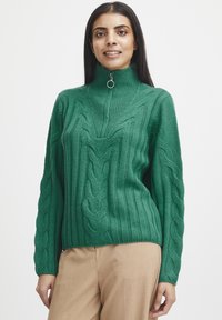 Grüner gerippter Strickpullover mit Kabelmustern, hohem Kragen und Viertelreißverschluss. Lange Ärmel mit strukturierten Details. Kombiniert mit beigen Hosen.
