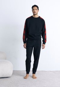 Schwarzes Sweatshirt und Hose Set, mit roten Logo-Akzenten an den Ärmeln, gerippten Bündchen und einem lockeren Schnitt. Weicher Stoff und glatte Textur.