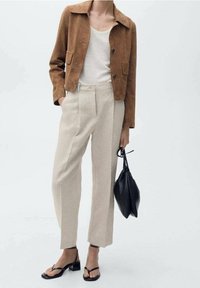 Bruine suède cropped jack, crèmekleurige tanktop, lichtbeige geplooide broek, zwarte sandaaltjes en een zwarte schoudertas.